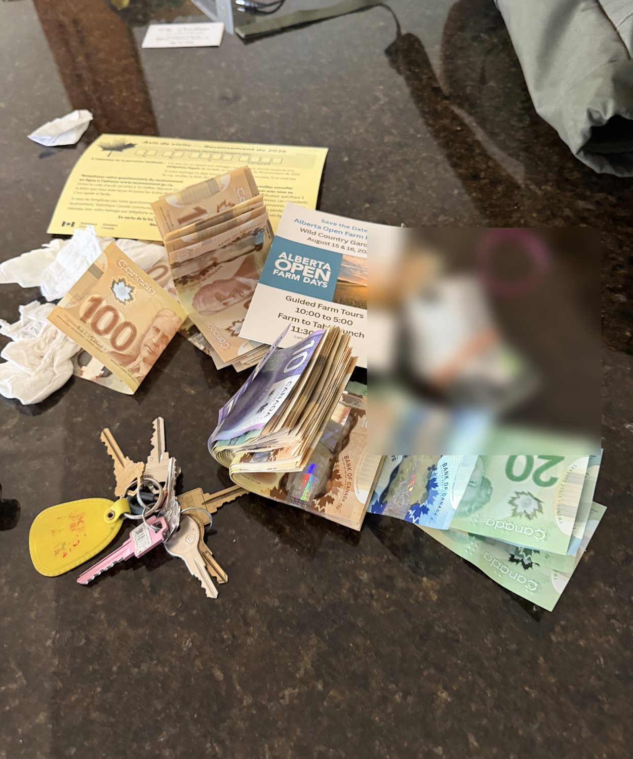 Tim's pocket contents on a granite counter: stacks of Canadian hundred-dollar bills, twenties and fives, a bundle of cards in an elastic band (blurred), a yellow keychain with keys, and an Alberta Open Farm Days flyer